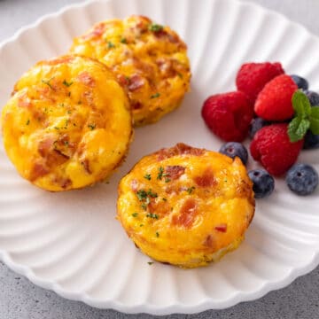 3 cheddar and bacon eggs bites garnished with snipped chives on a white plate. Served with berries on the side.