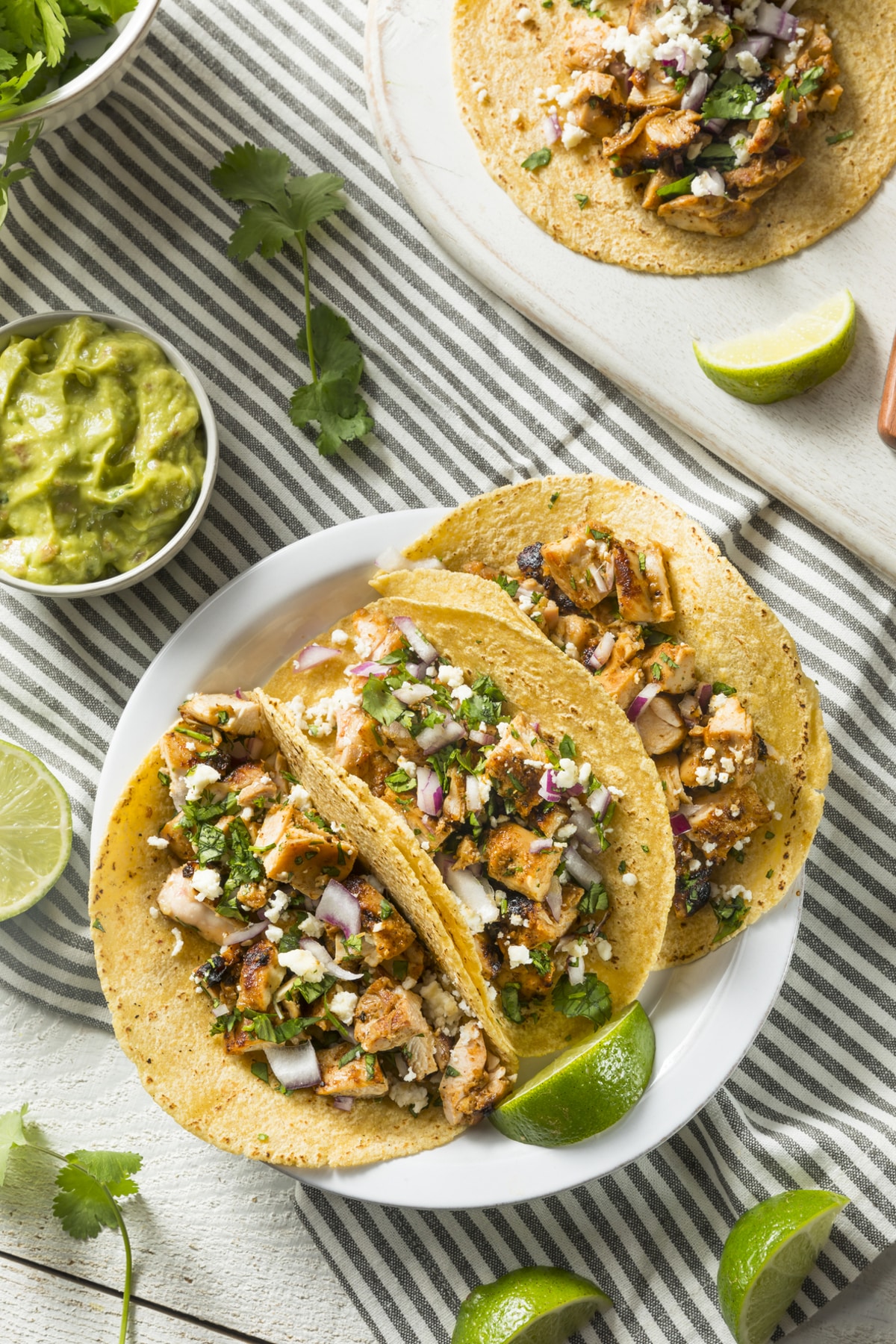 Chicken Tinga Tacos viewed from the top down. 3 tacos are assemble and on a plate. Guacamole and limes are served as garnishes on the side.