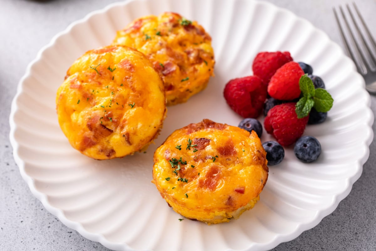 3 cheddar and bacon eggs bites garnished with snipped chives on a white plate. Served with berries on the side.