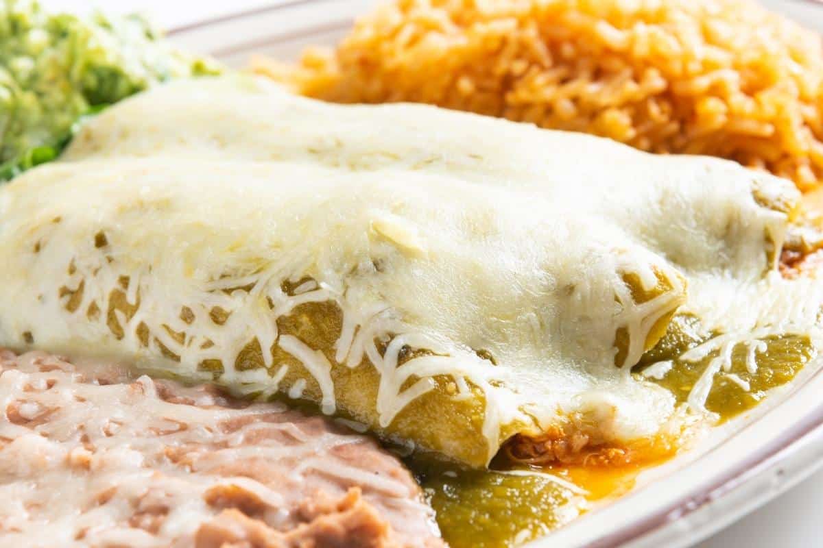 closeup of a plate loaded with green chili chicken enchiladas loaded with melted cheese and side servings of Mexican rice and refried beans.