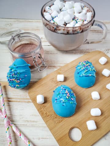 3 Easter blue chocolate bombs eggs sitting on a board with the hot chocolate bomb melted in a mug in the back.