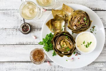 Instant Pot Artichokes trimmed and serve with dipping sauces and white wine. Top down view on a white plate and rustic white board